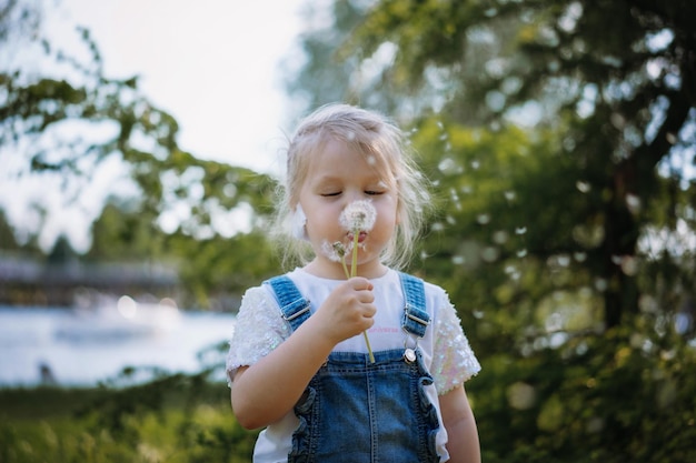 Pretty little girl blowing off dandelion seeds on sunset in summer park Image with selective focus