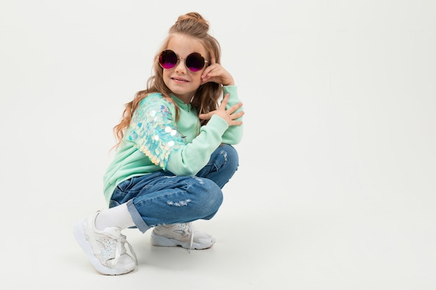 Pretty little girl in blue hoodie and sunglasses sits and smiles isolated on white