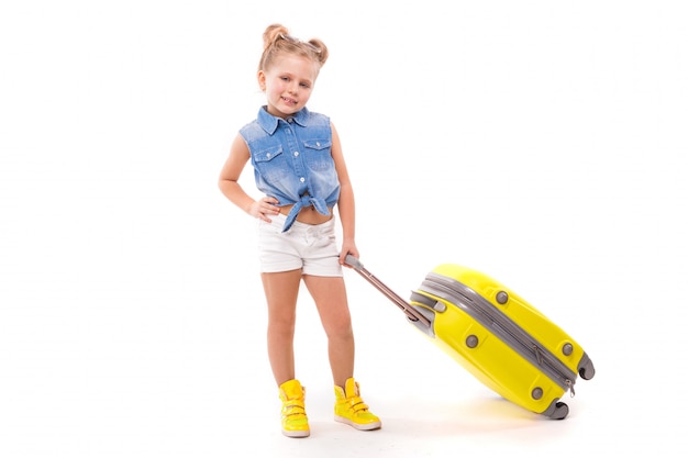 Pretty little girl in blue shirt, white shorts and sunglasses hold yellow suitcase by the handle