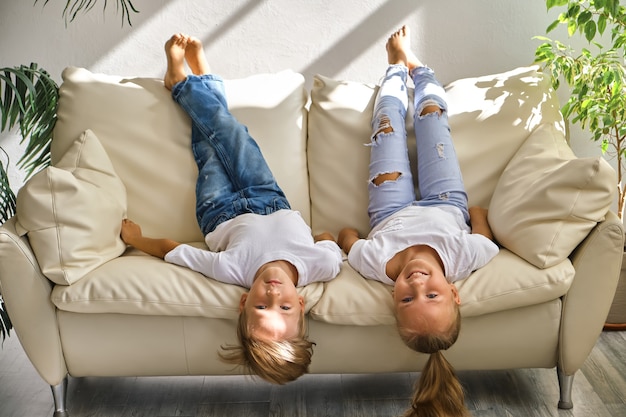 Pretty little girl and boy are lying on their backs on sofa in living room, looking at camera and smiling while playing at home