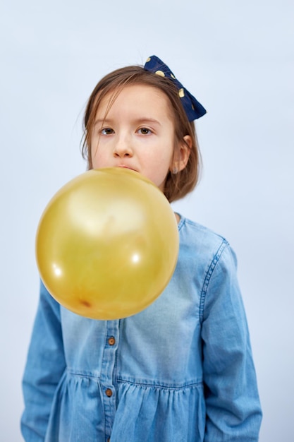 Pretty little girl in casual denim dress blowing inflate yellow balloon