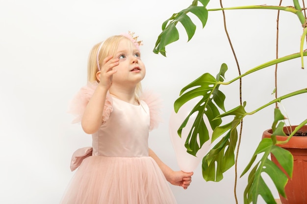 Pretty little girl in elegant pink dress in hands touches leaf of monstera houseplant On white background