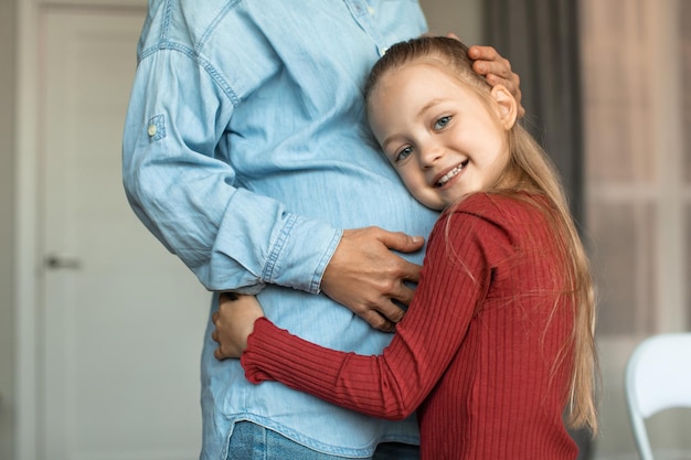 Photo pretty little girl hugging her pregnant mother big belly and smiling at camera kid bonding with expecting mom