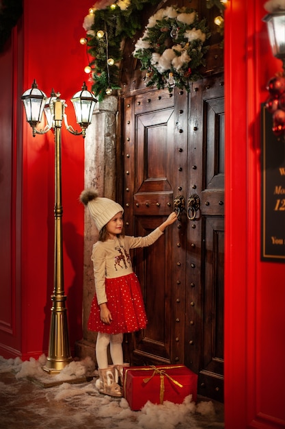 Pretty little girl is celebrating Christmasgiving red gift box Child knocking wooden door winter