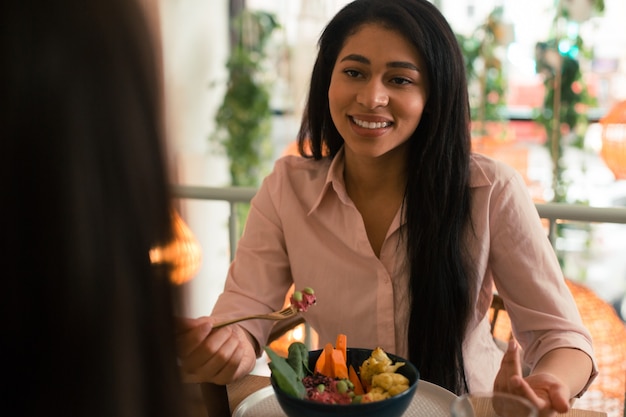 Pretty long haired lady smiling to her friend in a cafe while sitting with a bowl of vegetables