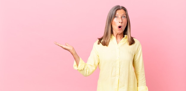 Pretty middle age woman looking surprised and shocked, with jaw dropped holding an object