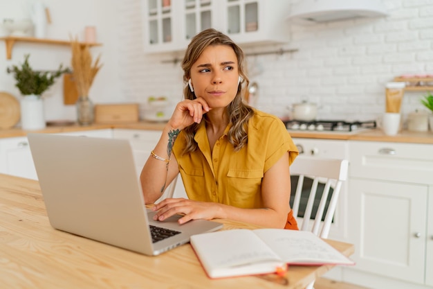 Pretty miling student woman using laptop and writing notes on her modern light kitchen