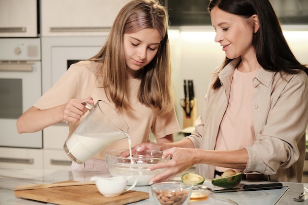 Pretty mother and her cute teenage daughter standing by table in the kitchen and pouring milk into bowl while prparing icecream