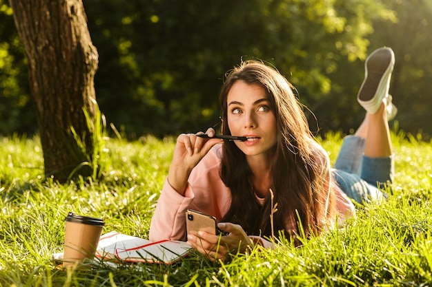 Pretty pensive young woman student using mobile phone while laying on a grass at the park