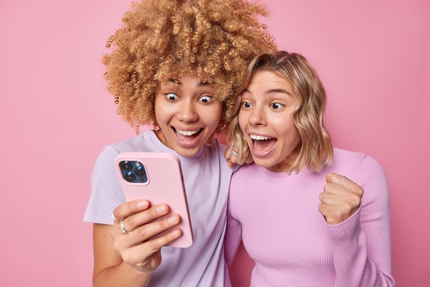 Pretty positive young womren stare happily at smartphone screen react on awesome news find out about winning of lottery exclaim from happiness isolated over pink background Wow thats great