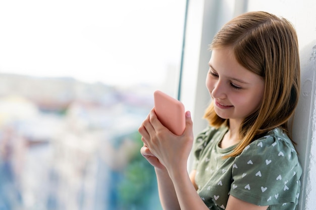 Pretty preteen girl looking at smartphone and smiling at home close to window with daylight beautifu