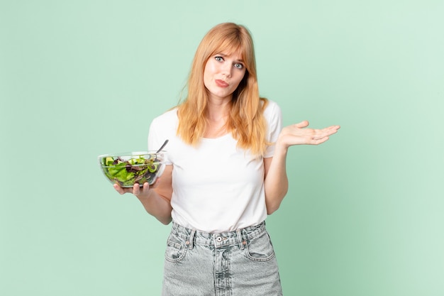 Photo pretty red head woman feeling puzzled and confused and doubting and holding a salad. diet concept