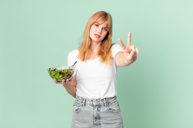 Photo pretty red head woman smiling and looking happy, gesturing victory or peace and holding a salad. diet concept