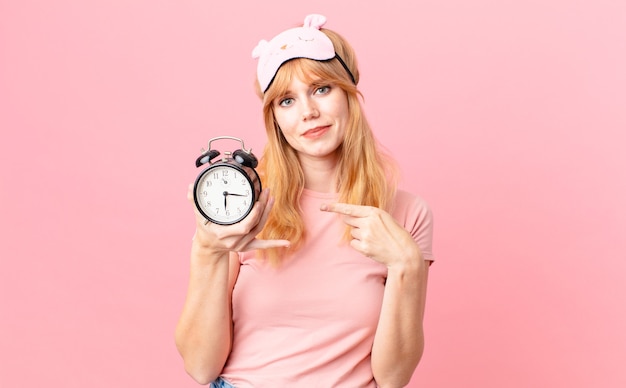 Pretty red head woman wearing pajamas and holding an alarm clock. wake up concept