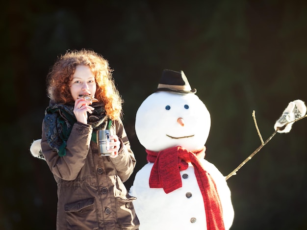 Pretty redhaired girl drinking tea and eating gingerbread outdoors with snowman at a winter day