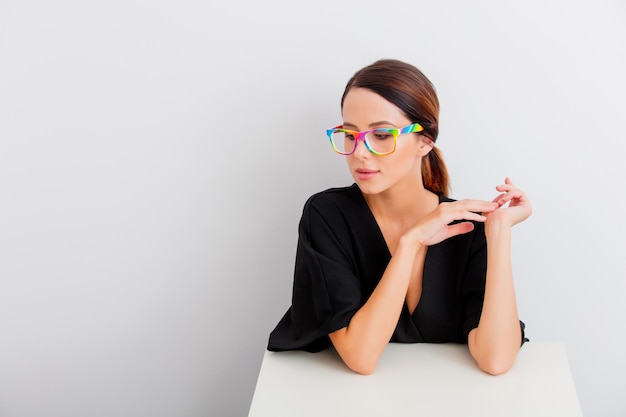 Pretty redhead caucasian woman in black dress and colored eyeglasses sitting at white table in lagom style on white background