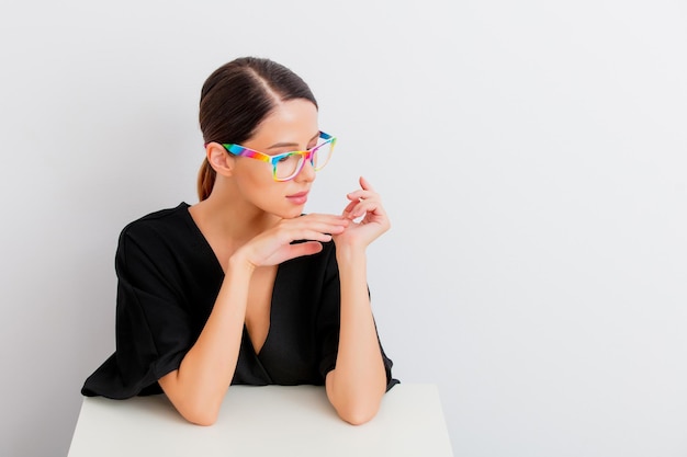 Pretty redhead caucasian woman in black dress and colored eyeglasses sitting at white table in lagom style on white background