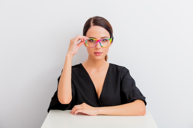 Pretty redhead caucasian woman in black dress and colored eyeglasses sitting at white table in lagom style on white background