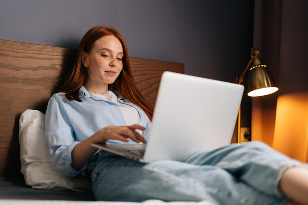 Pretty redhead young woman lying on bed and using computer laptop for using social media