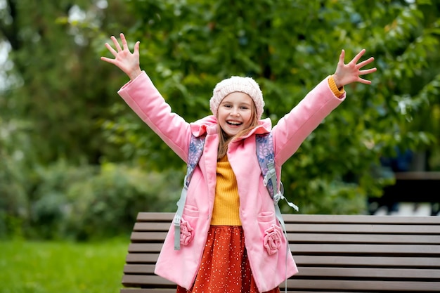 Pretty school girl kid with backpack looking at camera holding hands up and smiling at autumn park b