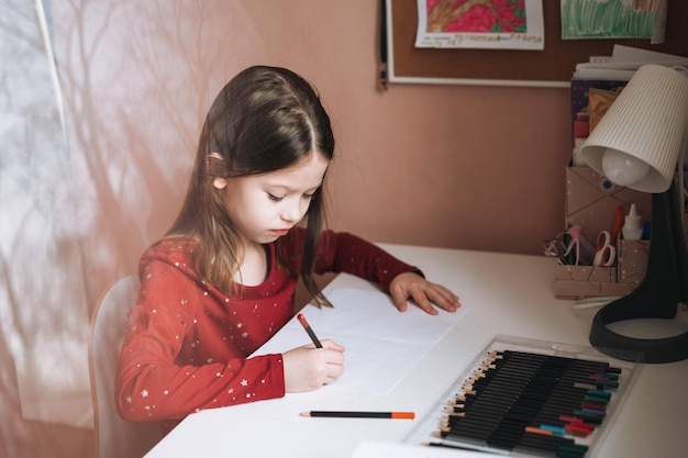 Pretty school girl with long hair in red dress draws at table in childrens room at home