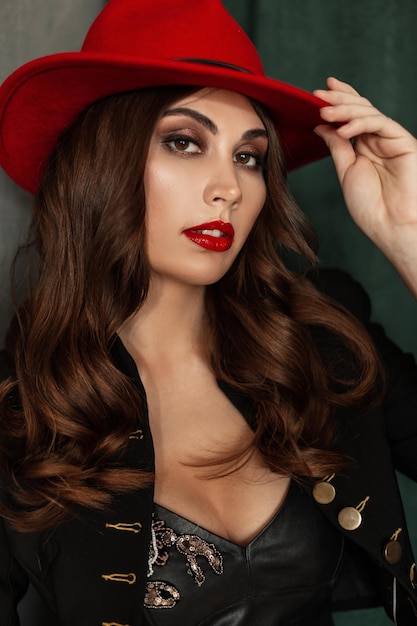 Pretty sexy woman model with makeup, red lips and curly hairstyle in fashion elegant clothes look with blazer and red hat poses near vintage background