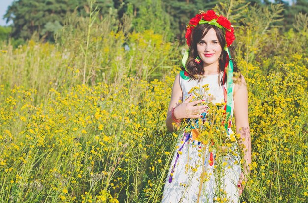 Pretty smiling girl in a garland of poppy flowers and national embroidered ethnic dress posing in the meadow of yellow field flowers lit with summer sun