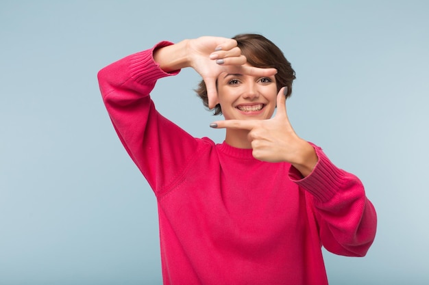 Pretty smiling girl in pink sweater making frame with fingers while happily looking in camera over blue background