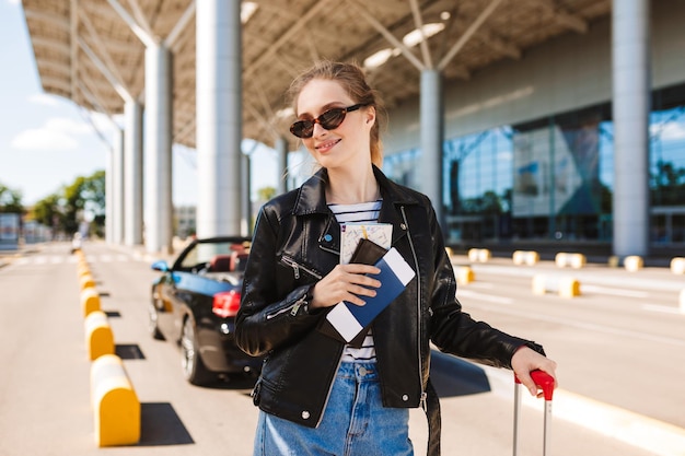 Pretty smiling girl in sunglasses and leather jacket with passport and flight ticket in hand joyfully looking in camera near airport with cabriolet car on background