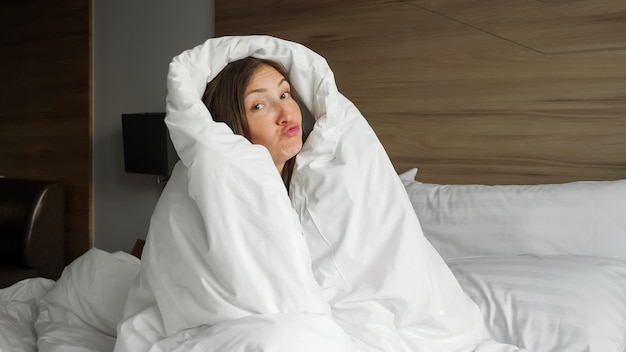 Pretty smiling girl in white blanket has fun in bed in bedroom in morning close-up