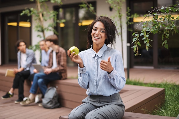 Pretty smiling lady sitting on bench with green apple and happily showing big thumb up gesture while 