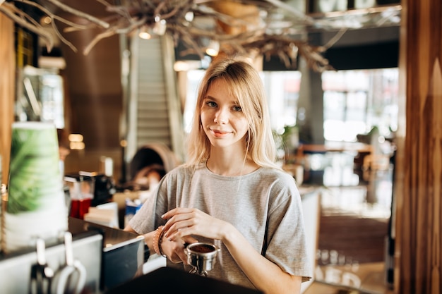 A pretty smiling slim girl,wearing casual clothes,holds some ground coffee and looks at the camera in a cozy coffee shop. .