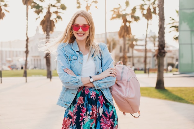 Pretty smiling woman walking in city street in stylish printed skirt and denim oversize jacket wearing pink sunglasses