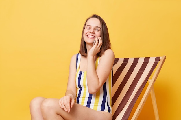 Pretty smiling woman wearing striped swimming suit sitting on deck chair isolated over yellow background talking on smart phone boasting her vacation