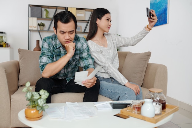 Pretty smiling young Asian woman taking selfie when her frowning husband looking at bills and pondering over household finances