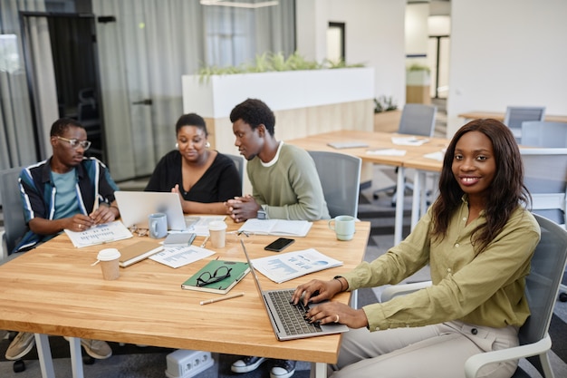 Pretty smiling young black businesswoman typing on laptop when attending business meeting with colle...