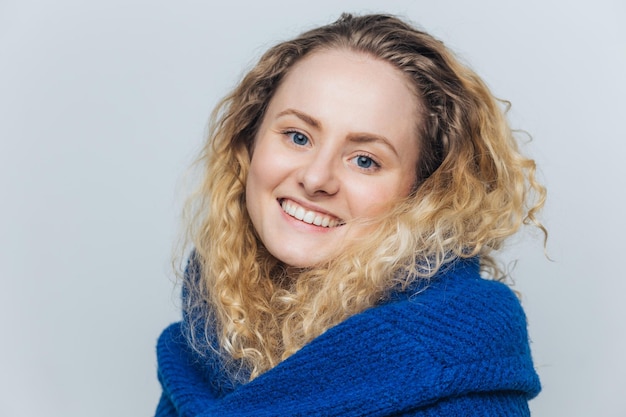 Pretty smiling young glad woman with blue warm eyes healthy skin and white perfect teeth dressed in warm sweater rejoices coming weekends isolated over light blue background models indoor