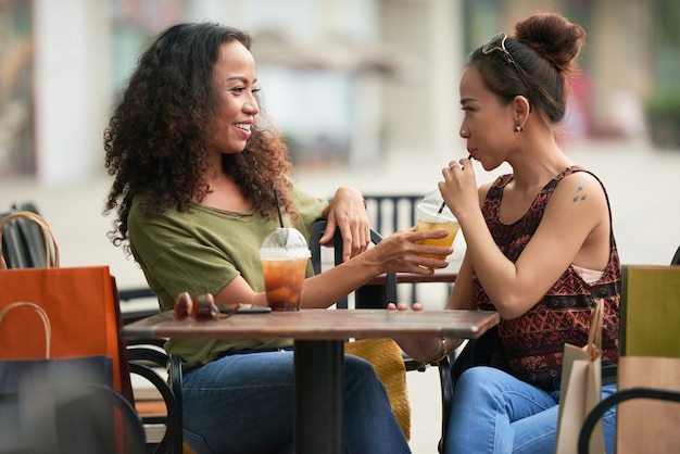 Pretty smiling young woman asking friend to taste her fruit cocktail when they are sitting at table in outdoor cafe