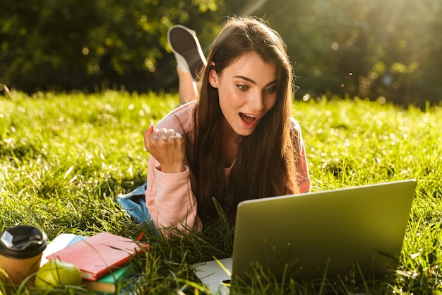 Pretty smiling young woman student studying on laptop computer while laying on a grass at the park, celebrating