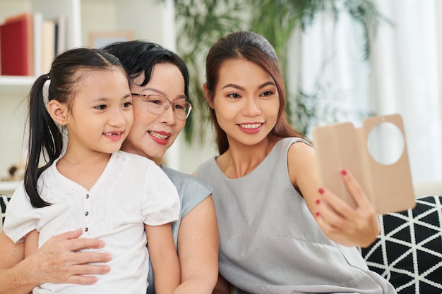 Pretty smiling young woman with smartphone taking selfie with her senior mother and little daughter