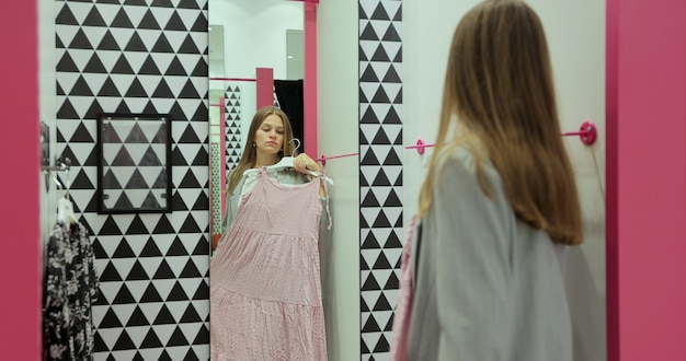 Photo pretty stylish caucasian girl trying on nice dress at mirror in dressing room in clothing shop. beautiful young woman choosing clothes in fancy sore of mall.