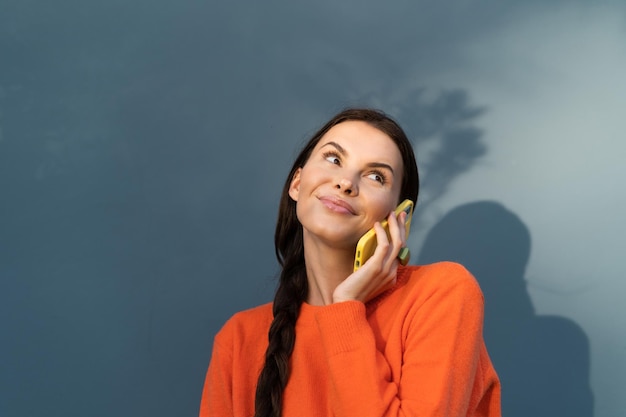 Pretty stylish woman in orange sweater posing on blue wall background outdoor cute making a call talking on mobile phone happy smiling