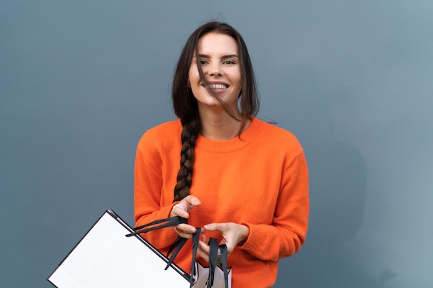 Pretty stylish woman in orange sweater posing on blue wall background outdoor wearing colorful rings accessories on fingers cute holding shopping bags big sale