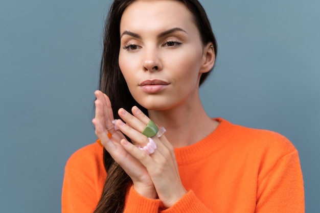 Pretty stylish woman in orange sweater posing on blue wall background outdoor wearing colorful rings accessories on fingers cute jewelry close shots