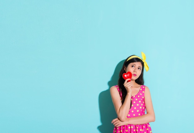 pretty sweet female student standing in blue wall background looking at empty area and holding heart daydreaming thinking future with cute dress.