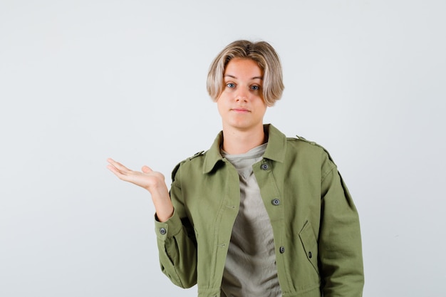 Pretty teen boy spreading palm aside in green jacket and looking indecisive , front view.