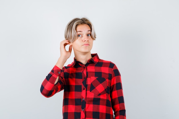 Pretty teen boy with hand behind ear in checked shirt and looking curious , front view.