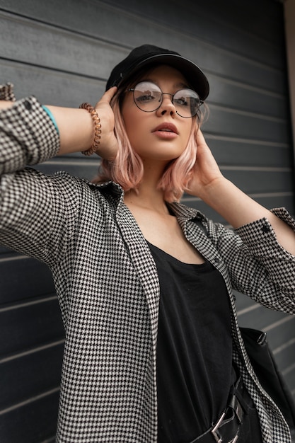 Pretty teenager model woman with pink hair and stylish glasses and a hat in fashionable casual youth clothing with a shirt and jeans posing near a gray gate on the street