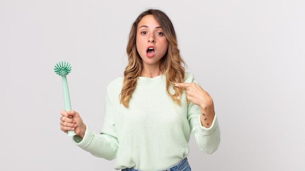 Pretty thin woman looking shocked and surprised with mouth wide open, pointing to self and holding a washing dish brush