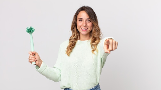 Pretty thin woman pointing at camera choosing you and holding a washing dish brush